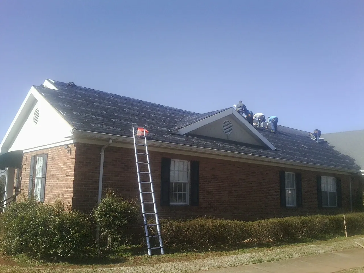 Crew tearing shingles off a brick ranch roof with a ladder set — Metal Roof Repair work by our crew in Pratt