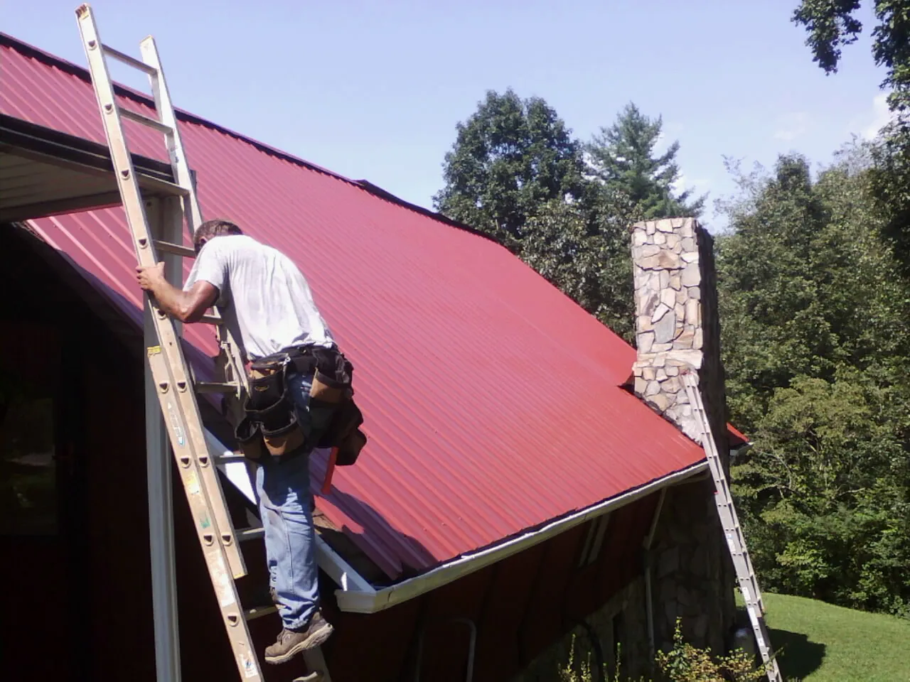 Roofer on a ladder beside a red metal roof and stone chimney — Soffit & Fascia Repair by Roofing Brothers in Pratt
